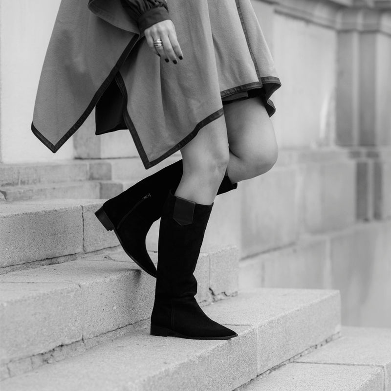 Mujer bajando unos escalones calzando unas botas planas de caña alta hechas en ante color negro