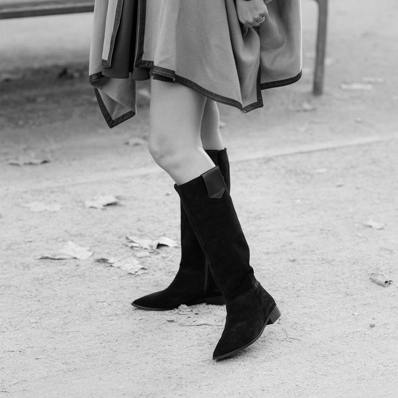 Mujer posando con un par de botas planas de caña alta clásica y elegante en ante negro.